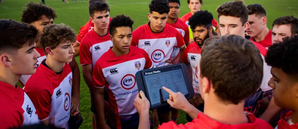 Heading: A group of rugby players standing around the coach wearing red Primal Media shirts. The coach is looking at a tablet with software displayed on it.